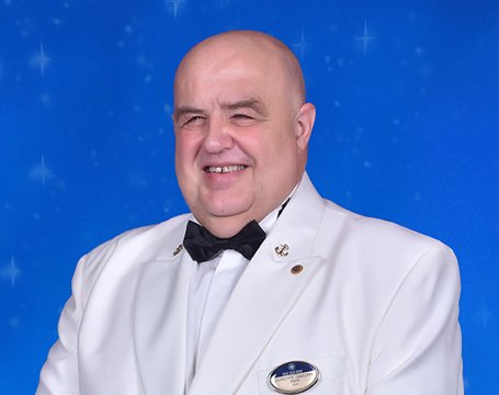 A crew member in a white uniform with an MSC badge against a blue starry background | MSC Cruises A crew member in a white uniform with an MSC badge against a blue starry background | MSC Cruises