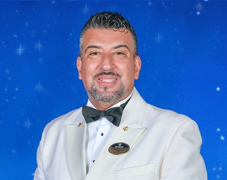 A crew member in a white formal uniform with an MSC name badge against a starry blue background | MSC Cruises A crew member in a white formal uniform with an MSC name badge against a starry blue background | MSC Cruises