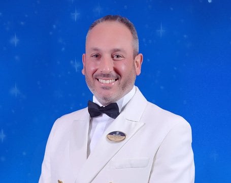 A crew member in a formal white uniform with an MSC Cruises badge against a starry blue background | MSC Cruises A crew member in a formal white uniform with an MSC Cruises badge against a starry blue background | MSC Cruises
